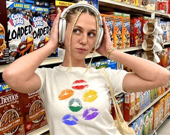 May include: A woman wearing a white t-shirt with a rainbow lip print and blue jeans is standing in a grocery store aisle. She is wearing headphones and holding a tote bag. The shelves are stocked with cereal boxes.