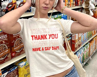 May include: A person wearing a white t-shirt with the text "Thank You Thank You Thank You Have A Gay Day!" in red letters. The person is wearing blue jeans and white headphones. The person is standing in a grocery store aisle with cereal boxes on the shelves.