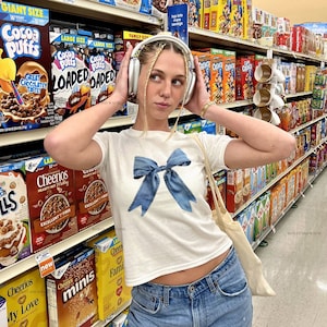 May include: A white t-shirt with a large blue bow design, paired with light-wash denim jeans. The person is wearing headphones and is in a grocery store aisle filled with cereal boxes.
