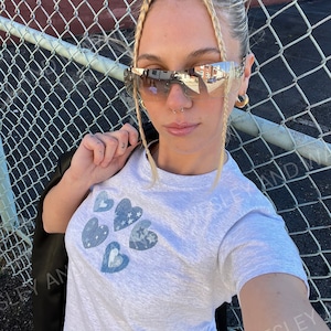 May include: A woman wearing a grey t-shirt with a blue heart design and blue jeans. She is wearing sunglasses and has her hair in a bun. She is standing in front of a chain link fence.