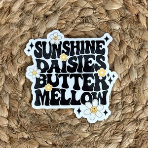 May include: A white sticker with black text that reads "Sunshine Daisies Butter Mellow" with a daisy graphic on each line. The sticker is on a woven brown background.