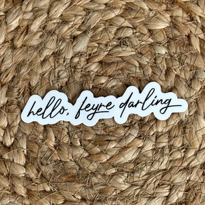 May include: A white sticker with the text "hello. feyre darling" in black cursive writing on a brown woven background.