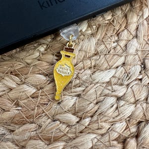 May include: A gold-colored charm in the shape of a potion bottle with the words "Felix Felicis" in yellow lettering. The charm is attached to a clear plastic clip and hangs near a black Kindle device.