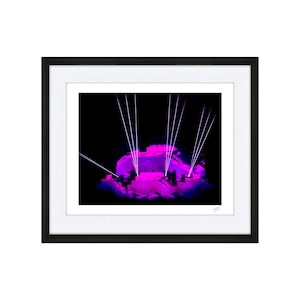Könnte beinhalten: Eine Bühne mit einer großen, rosa beleuchteten Struktur, auf die mehrere weiße Laserstrahlen scheinen. Die Bühne ist dunkel und die Struktur ist die einzige Lichtquelle.