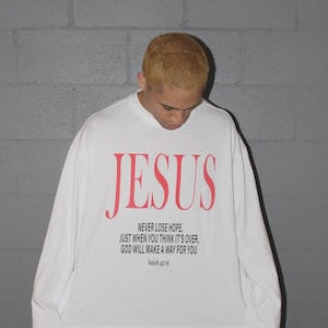 以下が含まれることがあります： 赤い文字で「JESUS」と書かれた白い長袖Tシャツ。下には「NEVER LOSE HOPE. JUST WHEN YOU THINK IT'S OVER, GOD WILL MAKE A WAY FOR YOU Isaiah 43:19.」と書かれています。