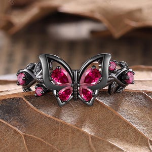 May include: A black butterfly ring with pink gemstones. The ring has a butterfly design in the centre, with pink gemstones set in the wings. Additional pink gemstones are set on the sides. The ring is displayed on a brown leaf.