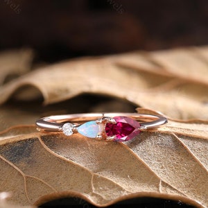 May include: A delicate rose gold ring with three pear-shaped gemstones. The ring features a ruby, an opal, and a small diamond.