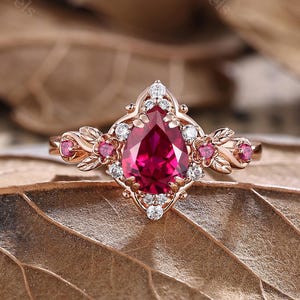 Peut inclure: Une bague en or rose ornée d'une pierre rouge foncé en forme de poire au centre, entourée de petites pierres roses et de pierres d'accent claires. La bague présente un design orné d'inspiration vintage avec des détails en forme de feuilles.
