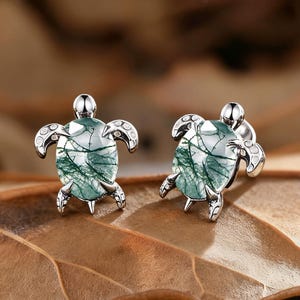 Peut inclure: Une paire de boucles d'oreilles à clous en forme de tortue argentée. Chaque boucle d'oreille présente un motif de tortue avec une coquille en pierre verte et blanche. Les boucles d'oreilles sont présentées sur une feuille brune.