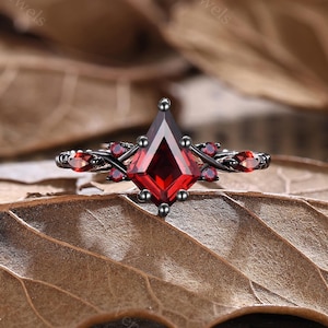 May include: A black ring with a large, kite-shaped, red gemstone as the centerpiece. Small red gemstones are set along the band and on the sides of the central stone. The ring is set against a backdrop of dried leaves.