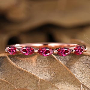 May include: A rose gold ring featuring a line of marquise-cut, vivid red gemstones. The ring is displayed on a bed of dried leaves, with the text "Venus VowJewels" in the background.