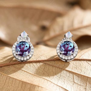 Può includere: Un paio di orecchini a lobo con una gemma rotonda e multicolore circondata da piccole pietre trasparenti. Gli orecchini hanno un design a alone e sono incastonati su un metallo dorato o argentato. Una pietra bianca più piccola è sopra.