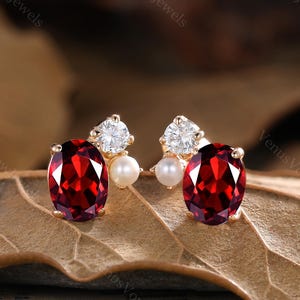 May include: A pair of gold stud earrings. Each earring has a large, oval-cut, deep red gemstone, a small, round pearl, and a cluster of sparkling white stones. The earrings are set on a brown surface.