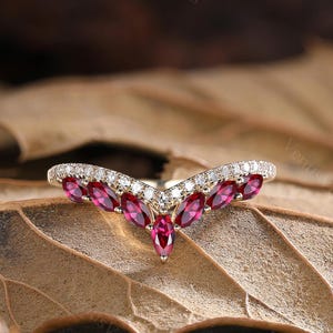 Peut inclure: Une bague dorée en forme de V. Elle présente une rangée de pierres précieuses rouges profondes taillées en marquise et de petites pierres précieuses claires. La bague est posée sur un fond de feuille brune.
