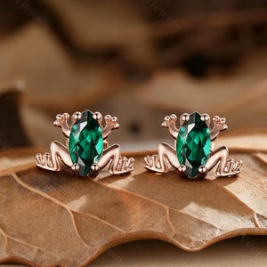 May include: A pair of rose gold frog-shaped stud earrings. Each earring has a marquise-cut, emerald-green gemstone set in the frog's body. The frog's legs and arms are detailed, and the earrings are displayed on a brown leaf.
