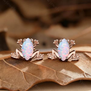 May include: A pair of rose gold frog-shaped stud earrings. Each earring features a light blue opal stone body with pink and white flecks. The frog's legs and arms are detailed, and the earrings are set on a brown leaf.