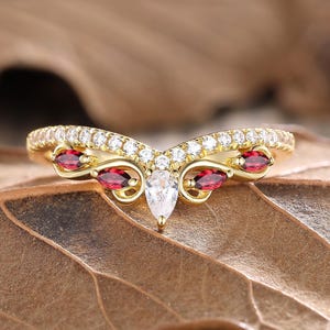 Peut inclure: Une bague dorée en forme de V. La bague est ornée de petites pierres rondes et transparentes et de pierres rouges ovales plus grandes. Une pierre transparente en forme de poire est centrée en bas du V. La bague est posée sur une feuille brune.