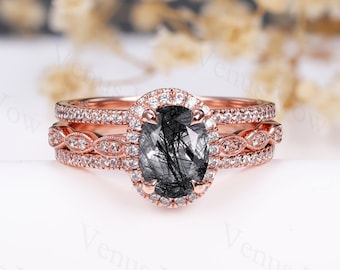 May include: A rose gold engagement ring set with a black and silver oval gemstone surrounded by a halo of smaller diamonds. The ring is part of a three-piece set, with a matching wedding band and a delicate band with a braided design.