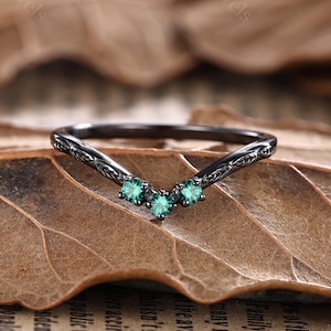 May include: A black V-shaped ring with three small, round, teal-colored gemstones. The ring has an ornate, etched design on the band. The ring is displayed on a brown leaf.