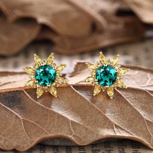 May include: A pair of gold-toned stud earrings. Each earring features a round, emerald-green gemstone at the center, surrounded by a halo of yellow gemstones. The earrings are set against a brown leaf.