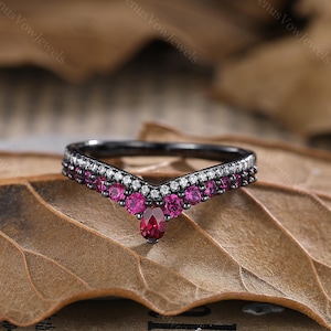 May include: Black band ring with a V-shaped design. The band is set with small clear and pink gemstones. The ring is on a brown leaf.