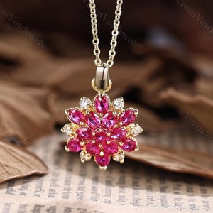 May include: A gold pendant necklace with a floral design. The pendant features a cluster of bright pink gemstones and small white stones, suspended from a fine chain. The necklace is set against a blurred background.