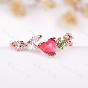 May include: A rose gold ring featuring a red heart-shaped stone, white cubic zirconia stones, and green stones. The ring has a delicate vine design.