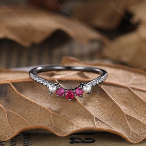Peut inclure: Bague noire avec une rangée de petites pierres claires et deux accents de perles. La bague a un design incurvé avec trois pierres roses au centre.