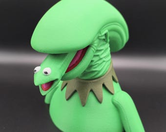 May include: Green 3D printed puppet head with a large, open mouth and googly eyes. The puppet is wearing a green collar with a brown trim.
