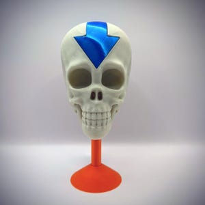May include: A white human skull replica featuring a prominent metallic blue arrow symbol on the forehead, mounted on a solid orange circular pedestal base. The skull has detailed eye sockets, a nasal cavity, and a full set of teeth in a neutral position.