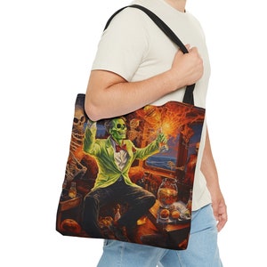 May include: A person is holding a black tote bag featuring a colourful print of a green-skinned skeleton in a suit, surrounded by other skeletons and flames. The bag has a black strap and is made of canvas.