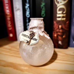 Può includere: Una bottiglia di vetro trasparente con un tappo bianco e gocciolante e un ciondolo a forma di chiave dorata. La bottiglia è piena di un liquido scintillante e ha un'etichetta che recita "VERITA-RUM".