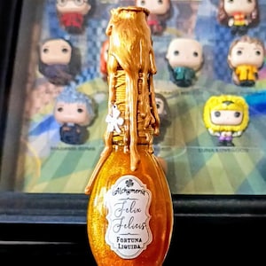 Può includere: Una bottiglia dorata con un'etichetta che recita "Alchymeria Felix Felicis Fortuna Liquida". La bottiglia è piena di un liquido dorato scintillante e ha un sigillo di cera dorato.