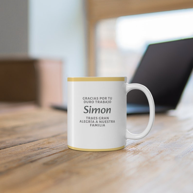 Peut inclure: Une tasse en c&eacute;ramique blanche avec un bord et une poign&eacute;e jaunes. La tasse porte l'inscription "Gracias por tu duro trabajo Simon Traes gran alegria a nuestra familia".