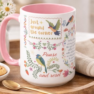 May include: A white ceramic mug with a pink interior and handle, featuring colorful illustrations of birds, flowers, and foliage. The text reads Just around the corner, Pause and reset, and a small section of scripture in a decorative floral frame.