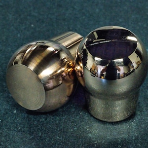 Puede incluir: Dos pomos de palanca de cambios metálicos con un acabado pulido y reflectante. Los pomos presentan una parte superior redondeada y esférica que se estrecha hacia una base cilíndrica. La superficie del metal muestra reflejos del entorno.