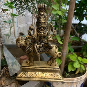 Peut inclure: Une statue en laiton détaillée d'une divinité chevauchant un lion. La statue est de couleur dorée et présente des sculptures complexes. La divinité a plusieurs bras et une coiffe détaillée. Le lion est également très détaillé, avec une crinière fluide et une forme musclée.