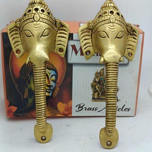 May include: Two brass elephant head door pulls. The elephant heads have detailed crowns and trunks that form the handle. The background includes a box with the words "Brass Articles" and images of religious figures.