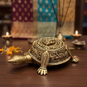 May include: A detailed brass turtle figurine with intricate patterns on its shell and body. The turtle is positioned on a wooden surface, with a blurred background featuring candles and decorative fabrics. The figurine is a decorative item.