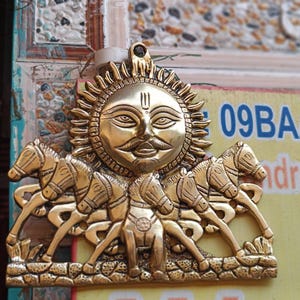 Könnte beinhalten: Ein goldfarbener Wandbehang aus Metall, der den Sonnengott Surya darstellt, der einen von vier Pferden gezogenen Wagen fährt. Der Sonnengott hat einen Bart und einen Schnurrbart und trägt eine Krone. Die Pferde sind in einer laufenden Position.