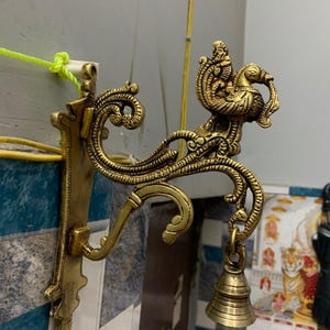 Peut inclure: Suspension murale en laiton orné avec un motif de paon. La pièce décorative comprend une cloche et des volutes complexes. Le support mural est fixé à un mur avec un motif de carreaux bleus et blancs.
