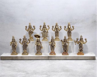 Könnte beinhalten: Eine Sammlung von zehn kleinen messingfarbenen Metallfiguren, die verschiedene Gottheiten darstellen und in zwei Reihen auf einem rechteckigen weißen Steinsockel angeordnet sind. Jede Figur zeigt feine Details und steht auf einem runden Sockel.