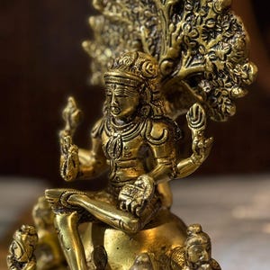 May include: A detailed golden statue of a seated deity with multiple arms, adorned with intricate jewellery and a headdress. The statue is surrounded by smaller figures and a decorative, tree-like element behind the head. The overall design is ornate and detailed.