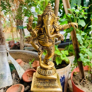 Könnte beinhalten: Eine goldfarbene Statue der hinduistischen Gottheit Ganesha, die auf einem dekorativen Sockel steht. Die Statue weist komplizierte Details auf, darunter mehrere Arme und einen Elefantenkopf. Der Hintergrund umfasst Topfpflanzen und Grün.