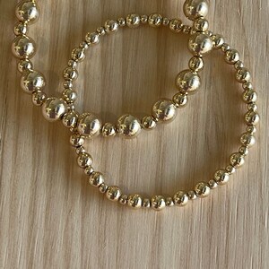 Könnte beinhalten: Zwei goldene Perlenarmbänder, eines größer als das andere, auf einer Holzoberfläche.