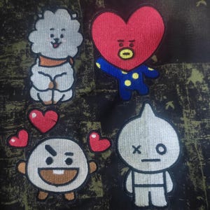 May include: Five embroidered patches featuring characters from the popular South Korean boy band BTS. The patches are on a black and green camouflage background. The characters are:  a white fluffy dog, a red heart with a face, a gray round character with a frown, a gray round character with an X and O for eyes, and a red heart.