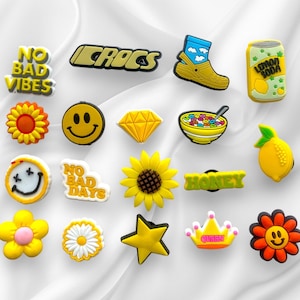 May include: A set of 16 yellow and orange shoe charms with various designs, including a smiley face, a sunflower, a lemon, a diamond, a bowl of cereal, a crown, a star, and the words "No Bad Vibes", "No Bad Days", "Crocs", and "Honey".