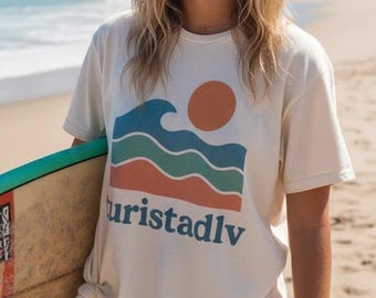 Könnte beinhalten: Cremefarbenes T-Shirt mit einem Wellen- und Sonnen-Grafik, Text "Turistadlv". Die Person hält ein Surfbrett an einem Sandstrand. Das Shirt wird mit braunen Shorts kombiniert.