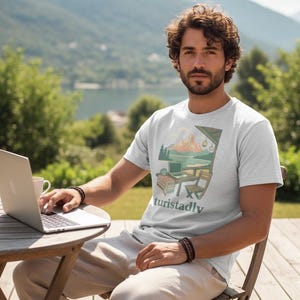 May include: Light gray t-shirt with a graphic of a mountain scene, laptop, and the word "turistadly" in teal. The shirt is being worn outdoors. A person is sitting at a wooden table with a laptop and a coffee cup.