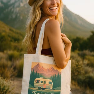 Puede incluir: Bolso tote color crema con una ilustración de caravana retro, montañas y árboles. El texto "Turistadly" está debajo de la caravana. Una persona con sombrero de paja lleva el bolso, sonriendo, con un fondo de montaña.